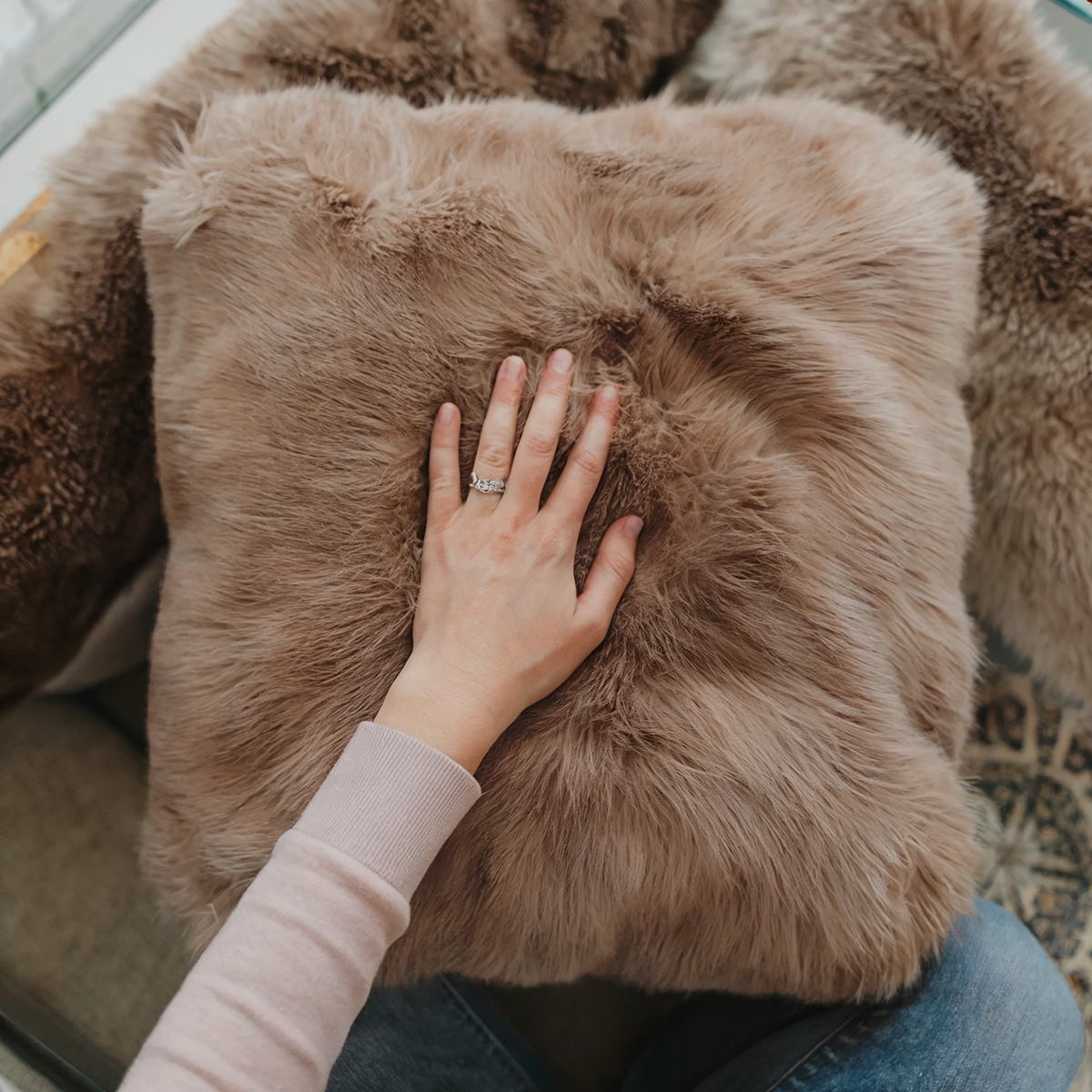 Close-up of a hand touching a taupe baby alpaca fur cushion, showing its soft texture and dense natural fiber